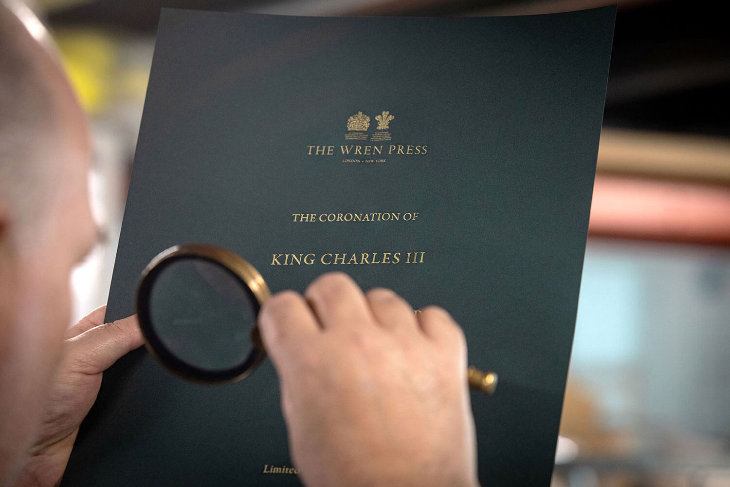 a man looking through a magnifying glass at die stamping work printed for the coronation of king charles by the wren press