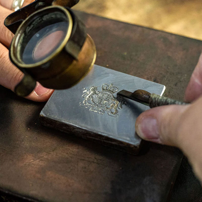 a master engraver carving a royal crest on a silver plate with tool and magnifying glass