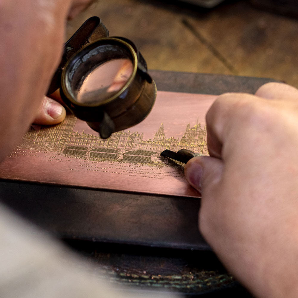 a master hand engraver working with a tool looking through a magnifying glass at a design on a copper plate